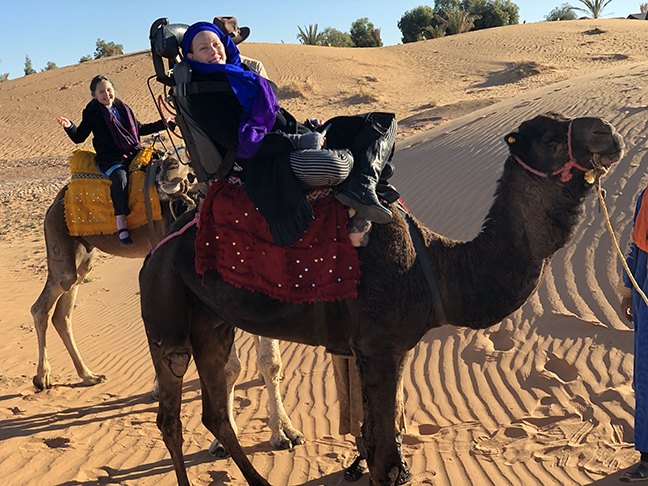 two camels in the desert, one in the foreground with a special accessible saddle and a woman on it, another in the background with a little girl on it
