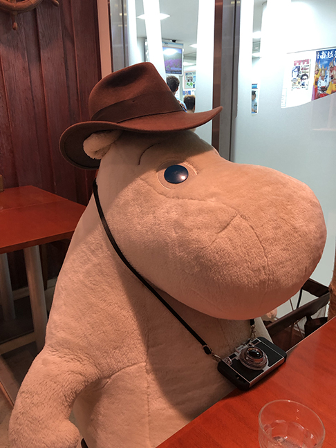 a giant stuffed Moomin stuffy with a brown fedora and camera on
