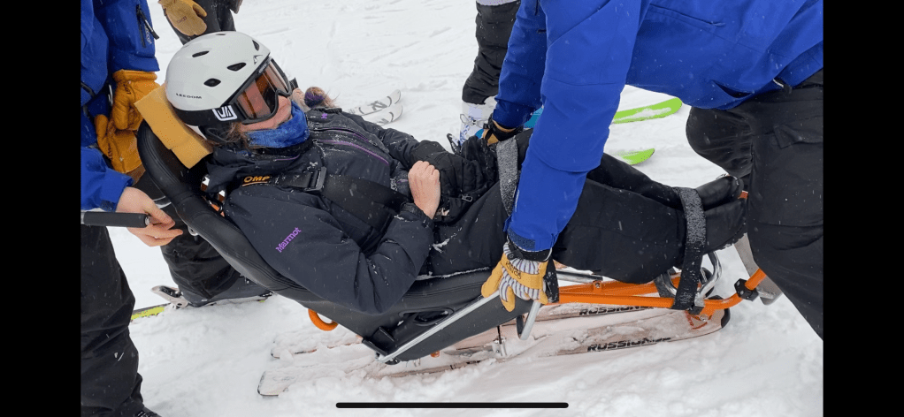 a woman with a helmet on laying in a reclining chair on skis.  She is strapped down.
