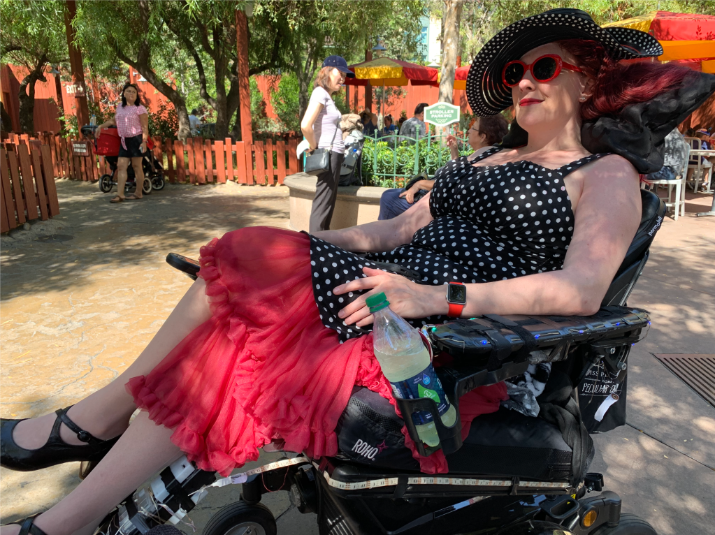 a woman in a black dress with white polka dots and a red petticoat, red sunglasses, red watch and black sun hat reclining in a wheelchair.