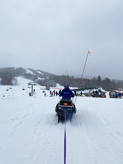 a snowmobile towing the person taking the picture by a long piece of purple webbing with a snow covered ski slope in the background