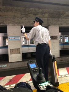 a man in a conductor's cap wearing white gloves and carrying a wheelchair ramp at a subway platform signals the train 
