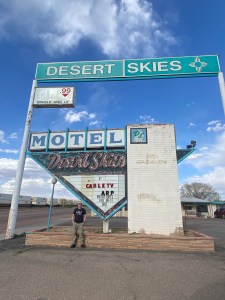 Man standing in front of an old faded neon sign which reads “Desert Skies Motel”