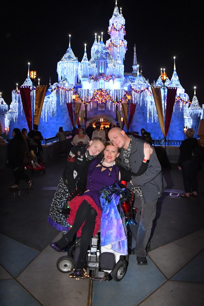 a family in front of the Disneyland castle at night.  The mother is in a wheelchair in the center and the daughter to the left and husband to the right.  They are hugging.