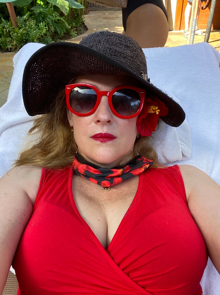 a woman (me) in a red shirt, black hat, red sunglasses and red lipstick close up, but obviously laying down.  Red hibiscus flower behind ear.