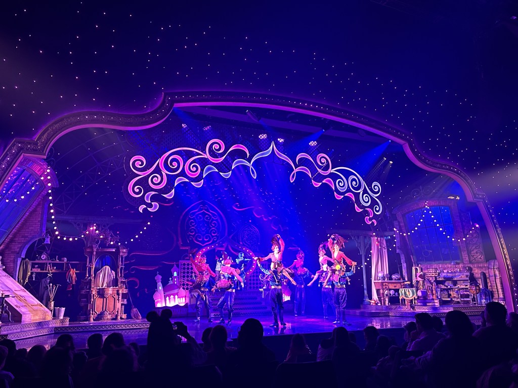 a dark stage show with stars, blue and pink lights, and dancers in pink and blue with the pink ladies on the men's shoulders