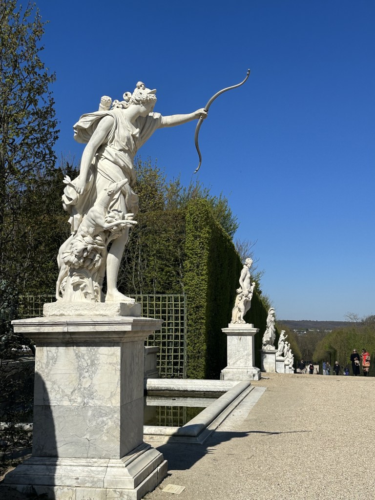 A series of greek statues in white marble on plinths receding into the distance in a garden with trimmed hedges.