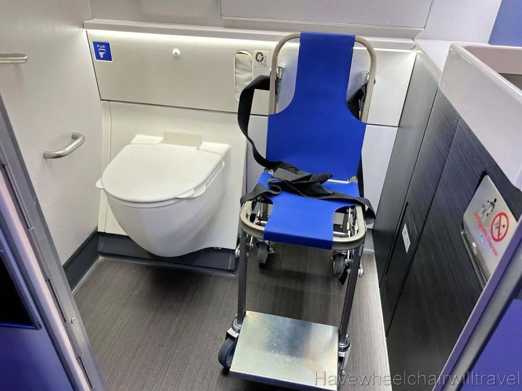 a picture of a large airplane bathroom with a toilet to the left and an aisle chair to the right with space in front and sink farther right.